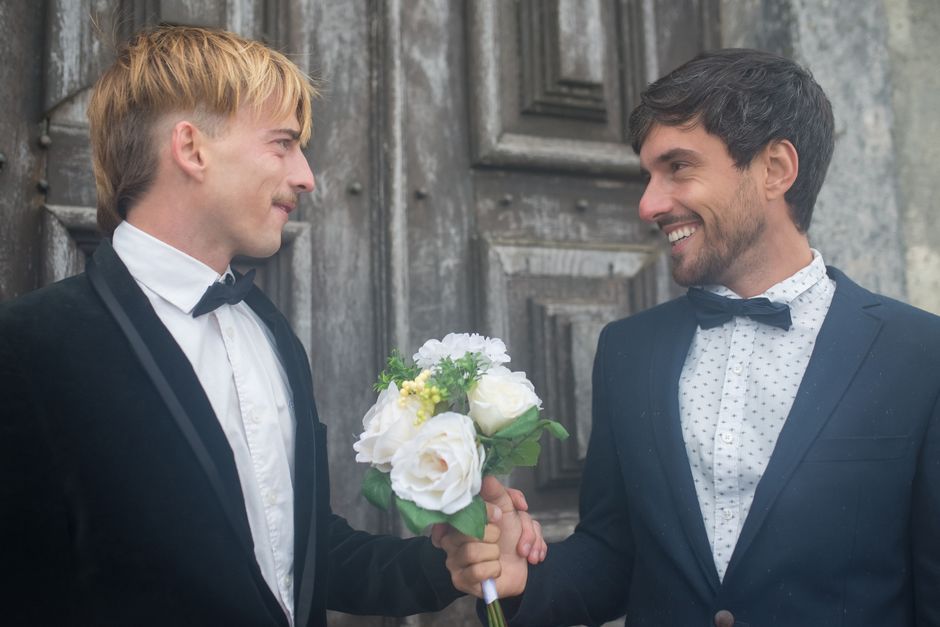 A man with dark hair and a beard wearing a navy blue suit with a white shirt and a black bow tie, smiling as he receives a floral bouquet from another man with blond hair dressed in a black tuxedo wit